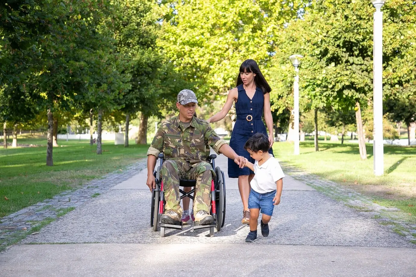 Ernst wirkender Militärvater, der im Rollstuhl mit seiner Familie unterwegs ist. Kaukasischer Vater mittleren Alters in Tarnuniform hält die Hand seines Sohnes und spricht mit seiner hübschen Ehefrau. Konzept von Kriegsveteran und Behinderung.